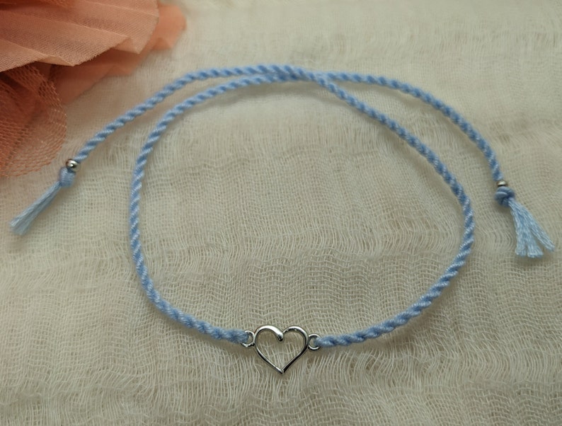 May include: A light blue friendship bracelet with a silver heart charm. The bracelet has two tassels on the ends.