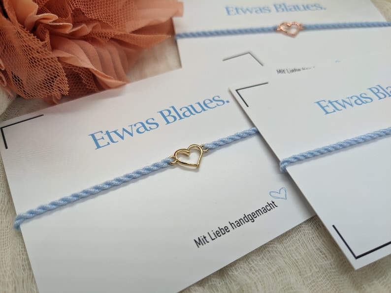 May include: A light blue bracelet with a gold heart charm. The bracelet is on a white card with the text "Etwas Blaues." and "Mit Liebe handgemacht".