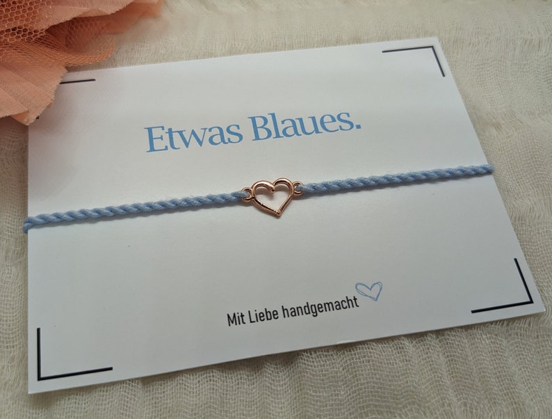 May include: A light blue bracelet with a rose gold heart charm. The bracelet is on a white card with the text "Etwas Blaues." and "Mit Liebe handgemacht".