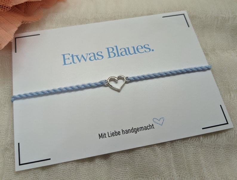 May include: A light blue bracelet with a silver heart charm. The bracelet is on a white card with the text "Etwas Blaues." and "Mit Liebe handgemacht".