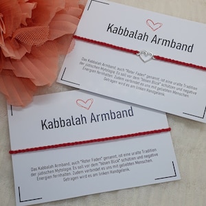 Könnte beinhalten: Zwei weiße Karten mit roten Schnurarmbändern und silbernen Herzanhängern. Die Karten tragen den Text "Kabbalah Armband" und eine Erklärung der Bedeutung des Armbands auf Deutsch.