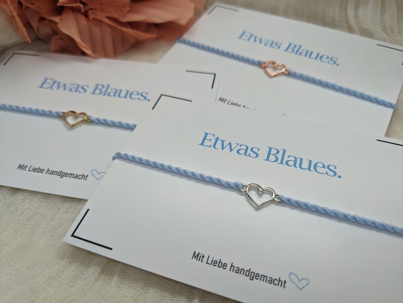 May include: Three cards with the text "Etwas Blaues." and the phrase "Mit Liebe handgemacht" on each card. Each card has a blue string bracelet with a heart charm.
