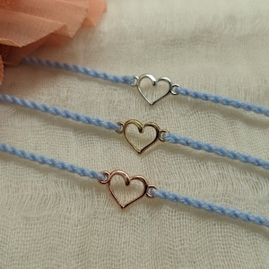 May include: Three delicate bracelets with a heart charm. The bracelets are made of a light blue cord and feature a silver, gold, and rose gold heart charm.