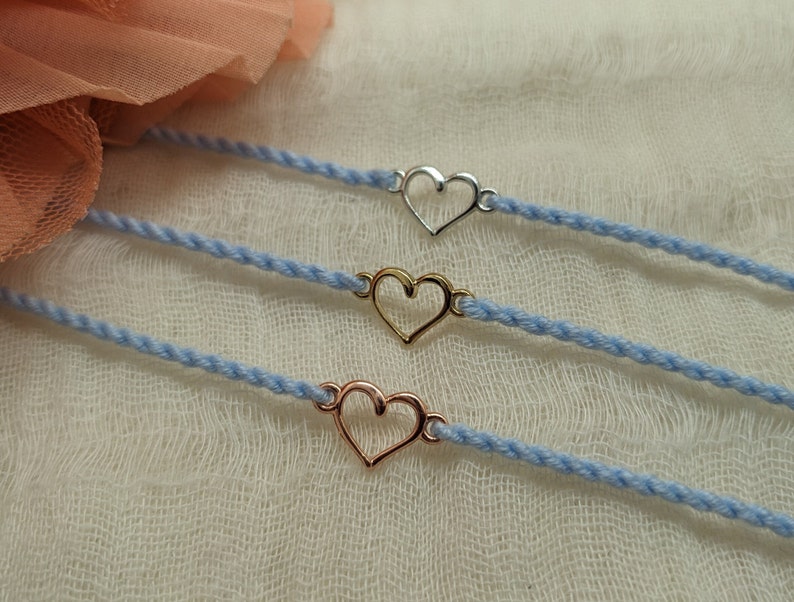May include: Three delicate bracelets with a heart charm. The bracelets are made of a light blue cord and feature a silver, gold, and rose gold heart charm.