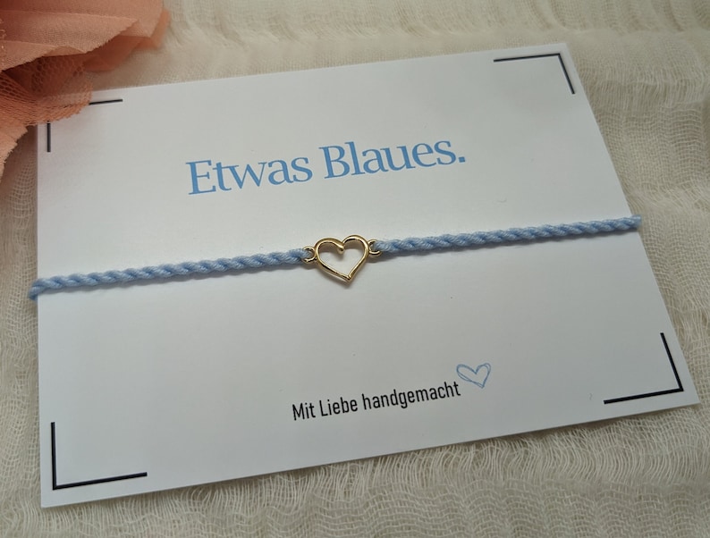 May include: A light blue bracelet with a gold heart charm. The bracelet is on a white card with the text "Etwas Blaues." and "Mit Liebe handgemacht".