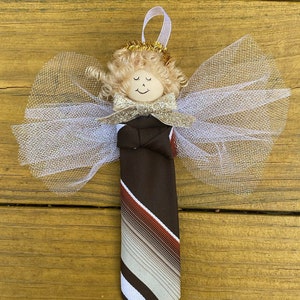 May include: A brown and white striped tie with a red stripe, tied into a bow and topped with a gold glitter bow. The tie is attached to a white tulle angel wing ornament with a blonde yarn doll head with a painted face and a gold glitter halo.