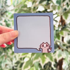 May include: A square, light blue sticky note with a brown border and a cartoon raccoon illustration in the bottom right corner. The raccoon is in a speech bubble. The background is a blurred green and white leafy plant.