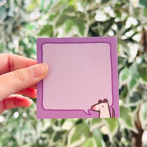 May include: A square, purple sticky note with a light purple interior and a dark purple border. A cartoon horse head is in the bottom right corner. The background is a blurred image of green and white leaves.