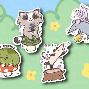 May include: Four colorful stickers featuring cartoon animals. A raccoon holding flowers sits on a mushroom, a frog with a grumpy expression sits on a red mushroom, a possum dances on a tree stump, and a mouse with wings holds a piece of cheese.