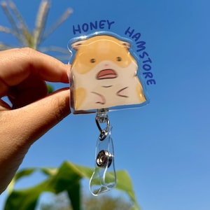 以下が含まれることがあります： 透明なHoney Hamstoreバッジリール。漫画のハムスターが描かれています。ハムスターは茶色と白で、「HONEY HAMSTORE」と青い文字で書かれています。リールには金属製のクリップと透明なプラスチックストラップが付いています。ハムスターは驚いた表情をしています。