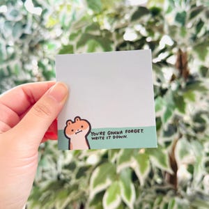 May include: A square sticky note with a light gray background and a green border. The border features a cartoon hamster and the text "YOU'RE GONNA FORGET. WRITE IT DOWN." The note is held by a hand.