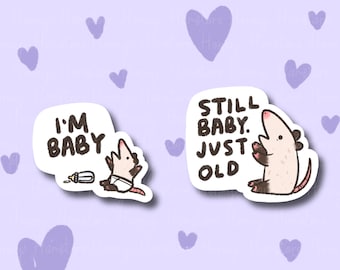 May include: Two stickers featuring opossum illustrations. One shows an opossum in a diaper with the text "I'M BABY" and a bottle. The other shows an opossum with the text "STILL BABY. JUST OLD". The background is light purple with lavender hearts.