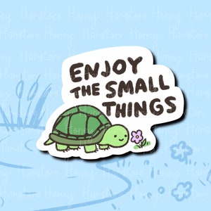 May include: A sticker featuring a cartoon turtle with a green shell and a smiling face, next to a small purple flower. The sticker has the text "ENJOY THE SMALL THINGS" in black letters on a white background.