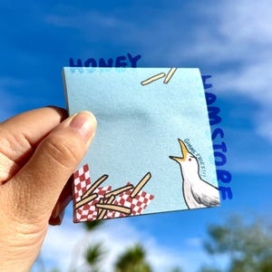 Peut inclure: Un post-it bleu clair avec le mot "HONEY" en bleu en haut. La note présente une mouette de dessin animé avec le texte "GIMME FRIES!!!" et un motif à carreaux rouges et blancs avec des frites.