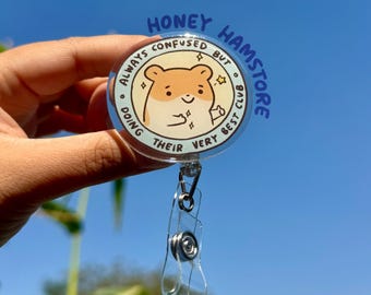May include: A clear, round retractable badge reel featuring a cartoon hamster. The hamster is light brown and white, with the text "ALWAYS CONFUSED BUT DOING THEIR VERY BEST CLUB" and "HONEY HAMSTORE" in blue.