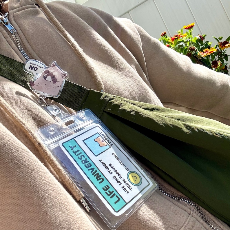 May include: A green and brown bag with a clear plastic ID card holder attached to it. The ID card has a blue background with white text that reads "Life University" and "Life Long Student." The card also has a small cartoon bear with a question mark above its head. The bag is hanging from a lanyard with a small raccoon charm.