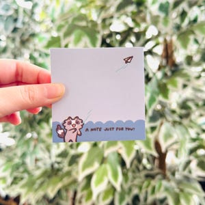 May include: A square, light blue sticky note with a cartoon illustration of a raccoon waving and a paper airplane. The text "A NOTE JUST FOR YOU!" is printed at the bottom. The background is a blurred view of green foliage.