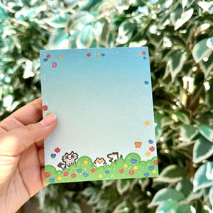 May include: A square notepad with a light blue gradient background. The bottom edge features a green grassy area with cartoon animals and colorful flowers. The top and sides are decorated with small flowers and butterflies. The notepad is held by a hand.