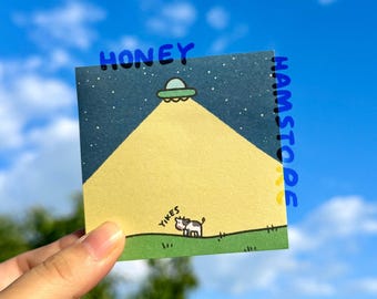 May include: A square sticker with a cartoon illustration of a UFO shining a light on a cow. The sticker has the words "HONEY" and "HAMSTODE" in blue, and "YIKES" near the cow. The background is a blue sky.