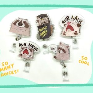 Op de afbeelding: Vijf verschillende kleurrijke plastic badgehouders met cartoon-wasbeerontwerpen. De ontwerpen omvatten een wasbeer met de tekst "Trash Gang", een wasbeer met de tekst "Aur Naur", een wasbeer met de tekst "Respectvol Nee", en twee wasberen met de tekst "Aur Naur".