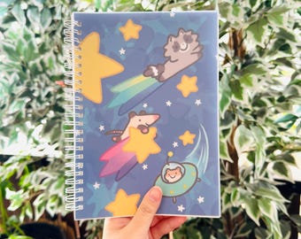Puede incluir: Un cuaderno con espiral con una cubierta azul que presenta ilustraciones de dibujos animados de un mapache, una zarigüeya y una criatura en un OVNI, todos montando estrellas fugaces. El diseño incluye estrellas amarillas y un fondo estrellado.