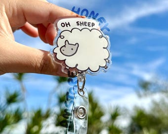 May include: A clear acrylic badge reel featuring a cartoon sheep design with the text "OH SHEEP". The sheep is white with a gray face and black outline. The reel has a clear plastic strap and a metal clip.
