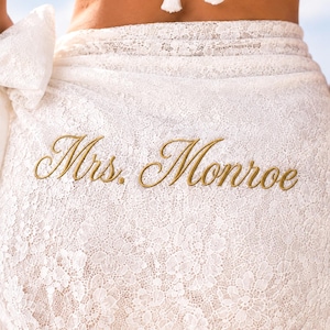 May include: A white lace sarong wrap featuring the embroidered text Mrs. Monroe in gold thread. The fabric has a detailed floral lace pattern throughout, with a tied knot detail on the side and white tassel accents visible at the top edge.