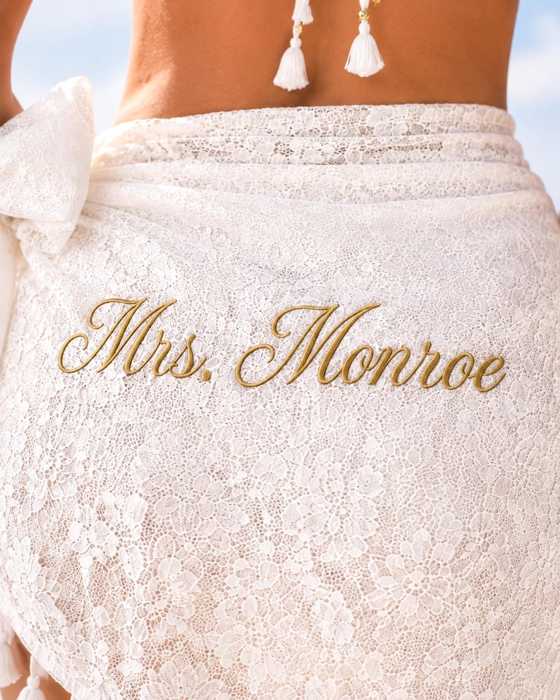 May include: A white lace sarong wrap featuring the embroidered text Mrs. Monroe in gold thread. The fabric has a detailed floral lace pattern throughout, with a tied knot detail on the side and white tassel accents visible at the top edge.