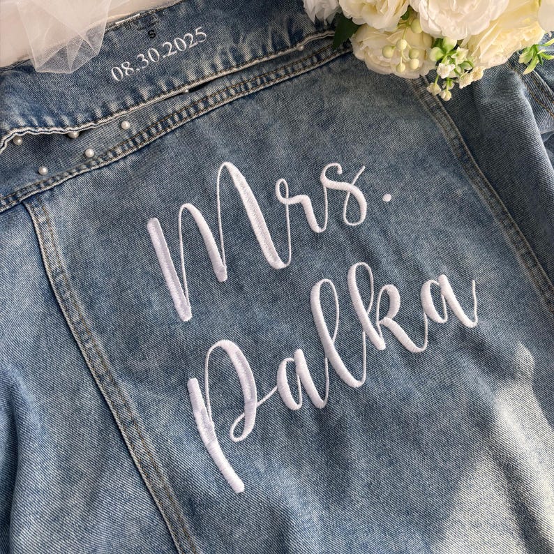 Puede incluir: Chaqueta vaquera azul claro con bordado blanco "Mrs. Palka" y la fecha "08.30.2025". La chaqueta tiene detalles de perlas y es talla S.