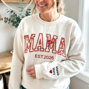 Könnte beinhalten: Cremefarbenes Sweatshirt mit dem Wort "MAMA" in rot geblümten Buchstaben. Der Text "EST.2026" steht unter dem Wort "MAMA". Der Ärmel trägt den Namen "Maisie Mary" und ein rotes Herz.