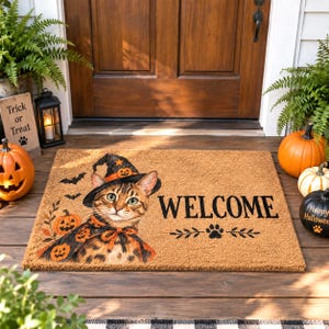 Peut inclure: Un paillasson rectangulaire en fibre de coco beige illustré d'un chat tigré brun portant un chapeau de sorcière noir et une cape assortie à motifs de citrouilles orange. Le mot WELCOME est inscrit en noir avec des chauves-souris et une patte.
