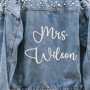 Puede incluir: Chaqueta vaquera azul claro con adornos de perlas en los hombros y el cuello. La parte trasera de la chaqueta presenta el texto bordado "Mrs. Wilson" en cursiva. Una elegante y personalizada chaqueta nupcial.