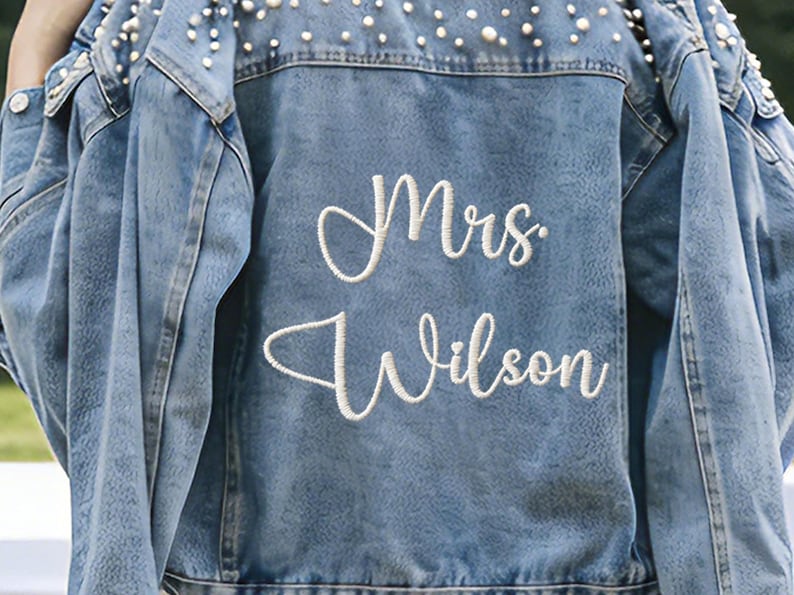 Puede incluir: Chaqueta vaquera azul claro con adornos de perlas en los hombros y el cuello. La parte trasera de la chaqueta presenta el texto bordado "Mrs. Wilson" en cursiva. Una elegante y personalizada chaqueta nupcial.
