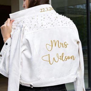 Puede incluir: Chaqueta vaquera blanca con adornos de perlas en los hombros. El texto bordado en dorado dice "Mrs. Wilson" y "22.3.20". La chaqueta tiene botones y un cuello cl&aacute;sico.