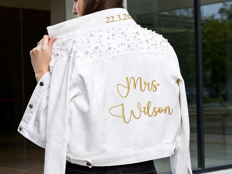 Puede incluir: Chaqueta vaquera blanca con adornos de perlas en los hombros. El texto bordado en dorado dice "Mrs. Wilson" y "22.3.20". La chaqueta tiene botones y un cuello cl&aacute;sico.