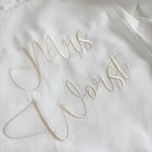 May include: A white fabric garment featuring the embroidered text "Mrs Vorst" in a cursive script. The embroidery is executed in a matching white thread, creating a subtle, tonal effect across the textured surface of the material.