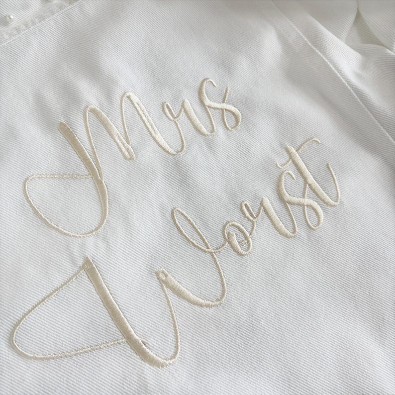 May include: A white fabric garment featuring the embroidered text "Mrs Vorst" in a cursive script. The embroidery is executed in a matching white thread, creating a subtle, tonal effect across the textured surface of the material.