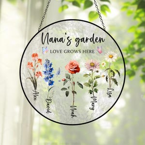May include: A round glass garden sign with a white background and a black border. The sign says "Nana's garden, Love Grows Here" with a butterfly on each side of the text. There are five flowers with names written below them: Alex, David, Noah, Mary, and Lisa.