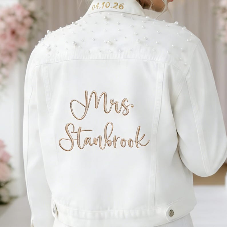 May include: A white denim jacket featuring scattered white pearls across the upper back and shoulders. The collar is embroidered with the date 04.10.26 in gold thread, while the back displays the embroidered gold text Mrs. Stanbrook in script.