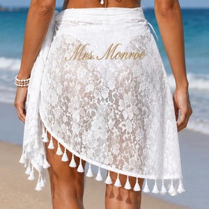 May include: A white lace sarong featuring a floral pattern and a hemline adorned with small white tassels. The fabric is semi-sheer, and the text Mrs. Monroe is printed in a gold script font across the center of the garment.