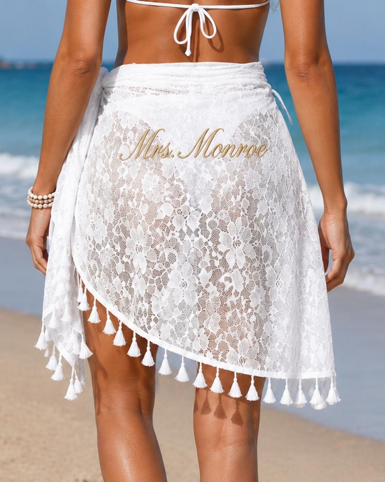 May include: A white lace sarong featuring a floral pattern and a hemline adorned with small white tassels. The fabric is semi-sheer, and the text Mrs. Monroe is printed in a gold script font across the center of the garment.