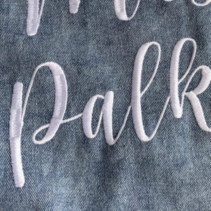 Puede incluir: Primer plano de tela vaquera con texto cursivo bordado en blanco que deletrea "Park". La costura es detallada y las letras est&aacute;n conectadas. El denim tiene un aspecto texturizado.