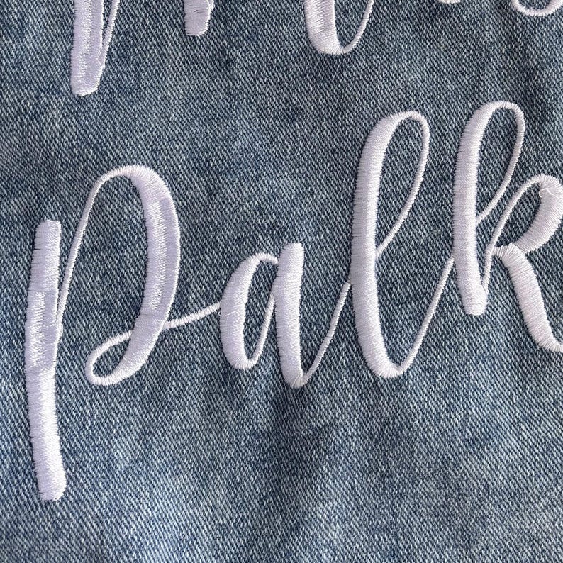 Puede incluir: Primer plano de tela vaquera con texto cursivo bordado en blanco que deletrea "Park". La costura es detallada y las letras est&aacute;n conectadas. El denim tiene un aspecto texturizado.