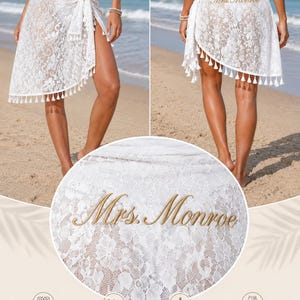 May include: A white lace sarong wrap featuring a delicate floral pattern, a tassel-trimmed hem, and gold embroidery on the back that reads "Mrs. Monroe." The garment is designed with an adjustable wrap fit for versatile styling over swimwear.