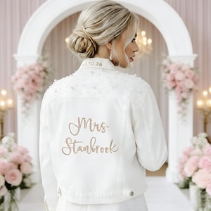 May include: White denim jacket with pearl embellishments and the embroidered text "Mrs. Stanbrook" in a cursive font. The jacket also features the date "04.10.26" embroidered on the collar. A bridal-themed jacket.