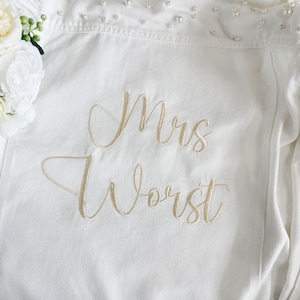 May include: A white denim jacket featuring the embroidered text "Mrs Worst" in a gold script font. The collar area is embellished with small white pearls, and a cluster of white artificial flowers is positioned on the left side of the garment.
