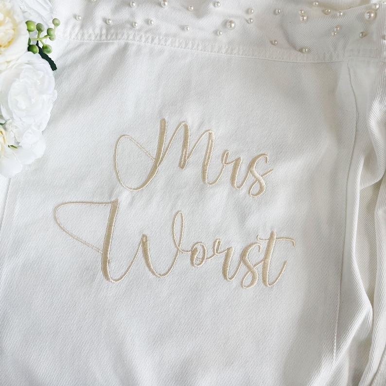 May include: A white denim jacket featuring the embroidered text "Mrs Worst" in a gold script font. The collar area is embellished with small white pearls, and a cluster of white artificial flowers is positioned on the left side of the garment.