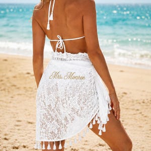 May include: A white lace sarong wrap featuring a floral pattern and fringe trim along the bottom edge. The fabric is embroidered with the gold text Mrs. Monroe. The garment is tied at the waist and worn over a white bikini top with thin straps.
