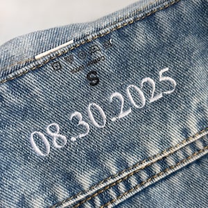 Puede incluir: Primer plano de un detalle de chaqueta vaquera azul claro. La chaqueta tiene costuras blancas y una fecha bordada en blanco, "08.30.2025." Se ve una peque&ntilde;a etiqueta negra de talla "S". La tela es 100% algod&oacute;n.