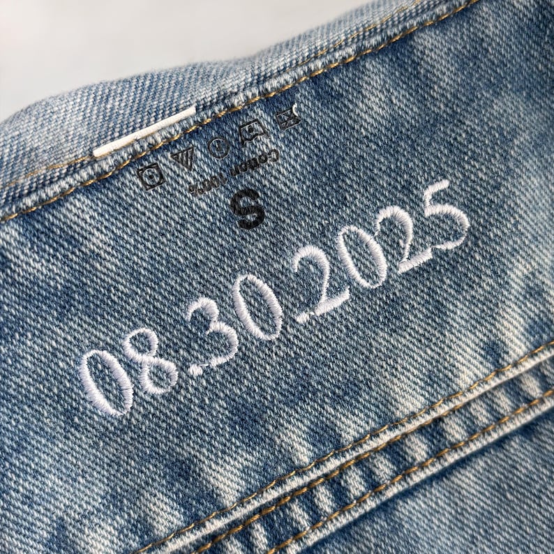 Puede incluir: Primer plano de un detalle de chaqueta vaquera azul claro. La chaqueta tiene costuras blancas y una fecha bordada en blanco, "08.30.2025." Se ve una peque&ntilde;a etiqueta negra de talla "S". La tela es 100% algod&oacute;n.